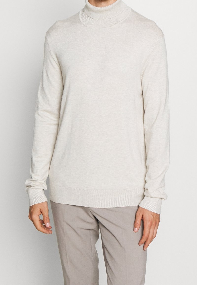 Ett krämfärgat polokrage-sweater gjort av mjukt stickat material. Har en figurnära design, ribbade manschetter och en slät textur. Bärs tillsammans med beige byxor.