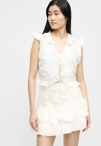 Joven con cabello largo y negro, vistiendo una blusa sin mangas blanca de encaje con volantes y una falda a juego de encaje, mirando hacia su izquierda.