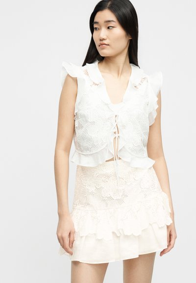 Νεαρή γυναίκα με μακριά μαύρα μαλλιά, που φοράει λευκό αμάνικο τοπ με δαντέλα και βολάν και matching φούστα από δαντέλα, κοιτάει αριστερά της.