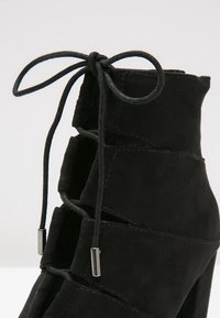 Bottine en daim noir avec devant à lacets ouverts et embouts de lacets argentés, montrant des coutures détaillées et une texture douce.