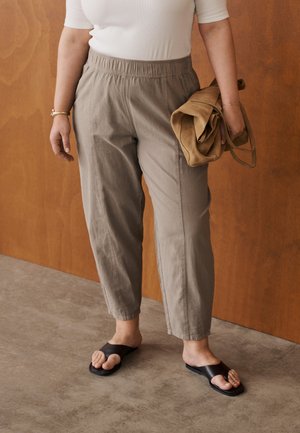 Osoba oblečená v svetlohnedých nohaviciach s elastickým pásom, bielom tričku, čiernych sandáloch s otvorenou špičkou, drží zloženú svetlohnedú tašku pred dreveným pozadím.