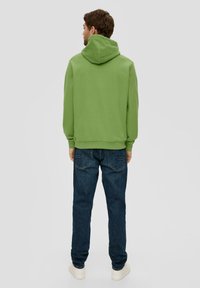 Sudadera verde con una gran capucha, corte relajado, puños acanalados, combinada con vaqueros azul oscuro y zapatillas blancas, vista desde atrás.