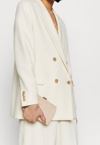 Bílý dvouřadý blejzr s velkými krémovými knoflíky, doplněný malým růžovým třpytivým psaníčkem. Hladká látka, ostré klopy a střih na míru.