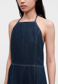 Femme portant une robe en jean bleu foncé à col halter avec des coutures blanches visibles sur un fond clair uni.