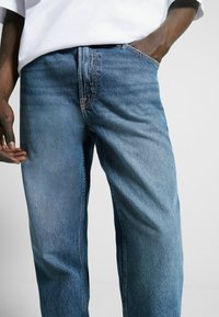 Jeans de mezclilla azul con un corte relajado, que presentan costuras visibles, bolsillos delanteros y un ligero desvanecimiento en el color. Combinados con una camiseta blanca.