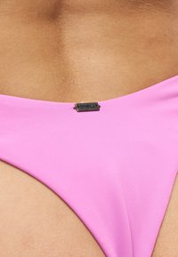 Ροζ κάτω μέρος μαγιό φορεμένο στη μέση ενός ατόμου με μια μικρή μεταλλική ετικέτα της μάρκας O'Neill τοποθετημένη στο κέντρο του υφάσματος.