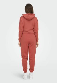 Hoodie u rust boji i odgovarajući trenerke od mekanog materijala, sa kapuljačom, elastičnim pojasom i rebrastim manžetama, upareni sa bijelim tenisicama.