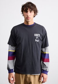 Tricou din bumbac negru cu textul "OBEY has a Posse". Mâneci gri cu dungi violet, roșu și galben. Croială lejeră.