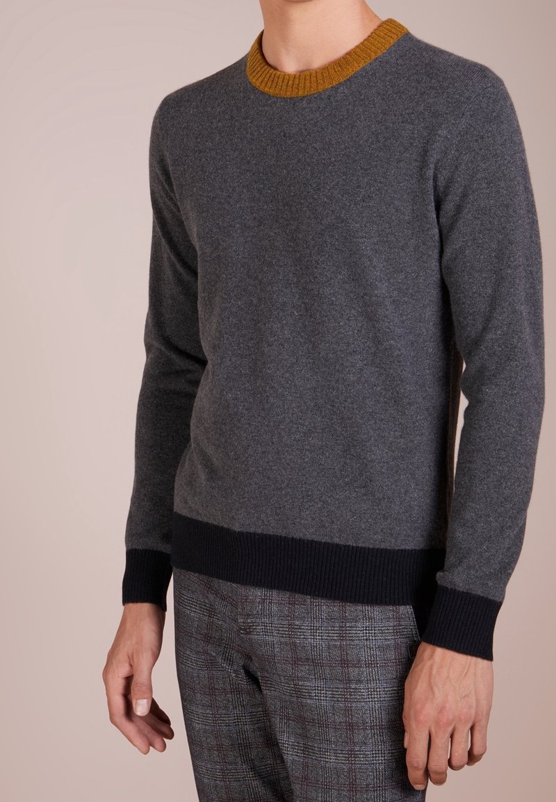 Homme portant un pull gris avec un col moutarde et des poignets noirs, associé à un pantalon à carreaux gris sur un fond uni.