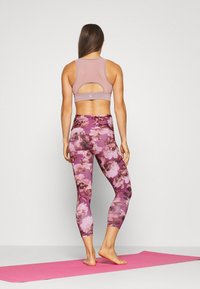 Kobieta stojąca boso na różowym macie do jogi, ubrana w mauve biustonosz sportowy i fioletowe legginsy w kwiatowy wzór, zwrócona plecami w kierunku jednolitego tła.