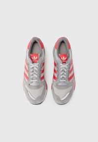 Adidas sneakers met een grijze mesh bovenkant, rode suede accenten, drie rode strepen en een witte zool met een reflecterende detail.