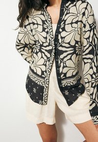 Czarna i kremowa koszula zapinana na guziki w kwiatowy wzór, wykonana z lekkiego materiału, z długimi rękawami, kołnierzykiem i ozdobnymi detalami.