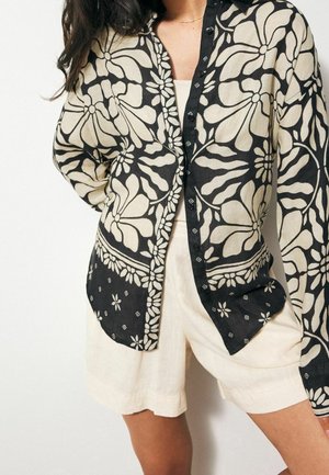 Chemise à boutons à motifs floraux noir et crème en tissu léger, avec des manches longues, un col, et des détails décoratifs.