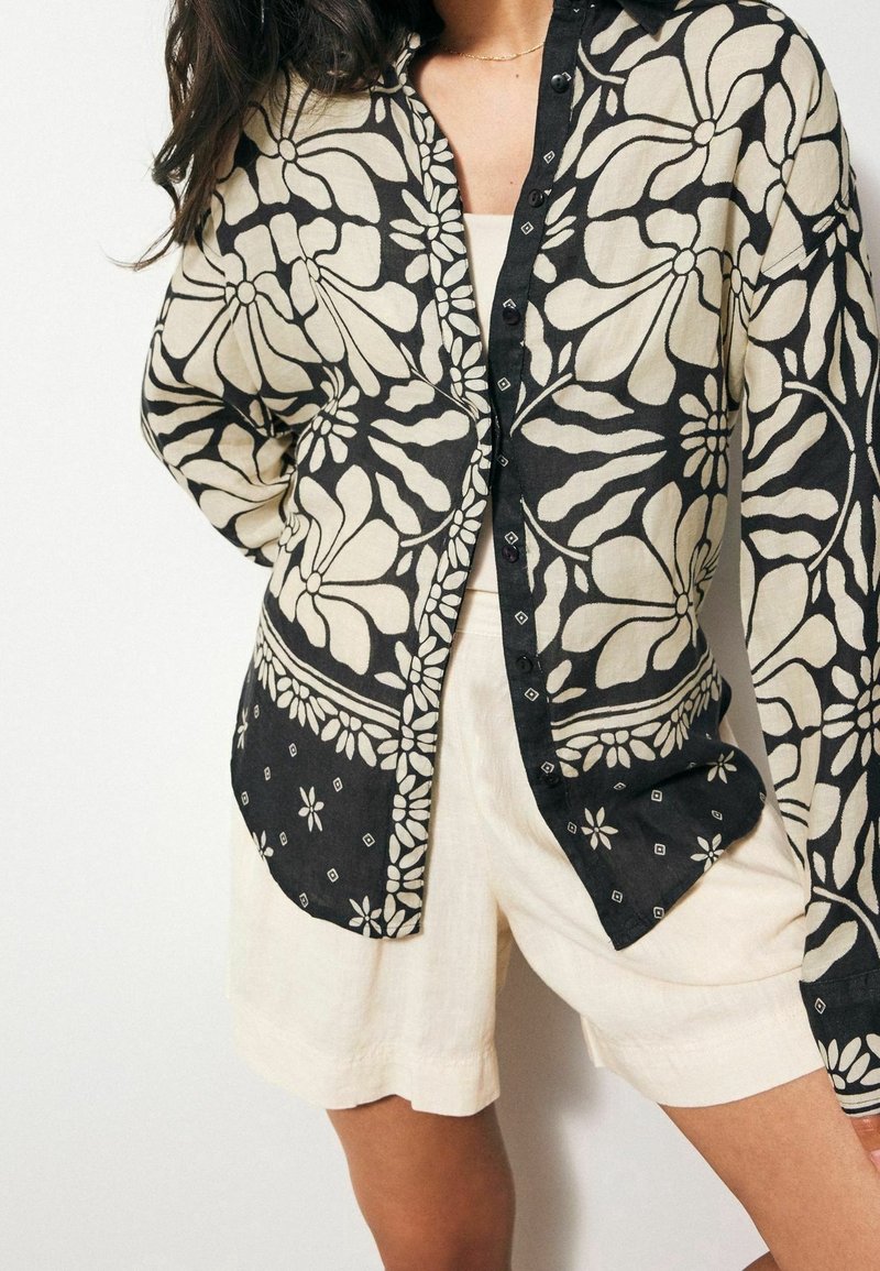 Czarna i kremowa koszula zapinana na guziki w kwiatowy wzór, wykonana z lekkiego materiału, z długimi rękawami, kołnierzykiem i ozdobnymi detalami.