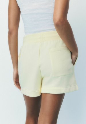 Isik kandmas helekollaseid kõrge vöökohaga lühikesi pükse, millel on tagatasku, ja valget varrukateta topi, taustaks lihtne hele taust.