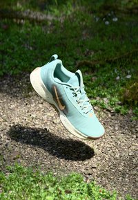 Baskets de sport bleu clair avec un tissu texturé, semelle blanche, accents dorés et un logo proéminent sur le côté, présentées sur un fond naturel.