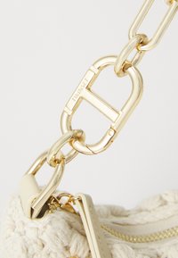 Gold metal chain attachment with a distinctive "T" logo, connecting to a cream textured fabric bag with a zipper detail.