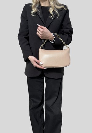 Donna con abito nero che tiene una borsa in pelle beige con tracolla a catena dorata su sfondo neutro