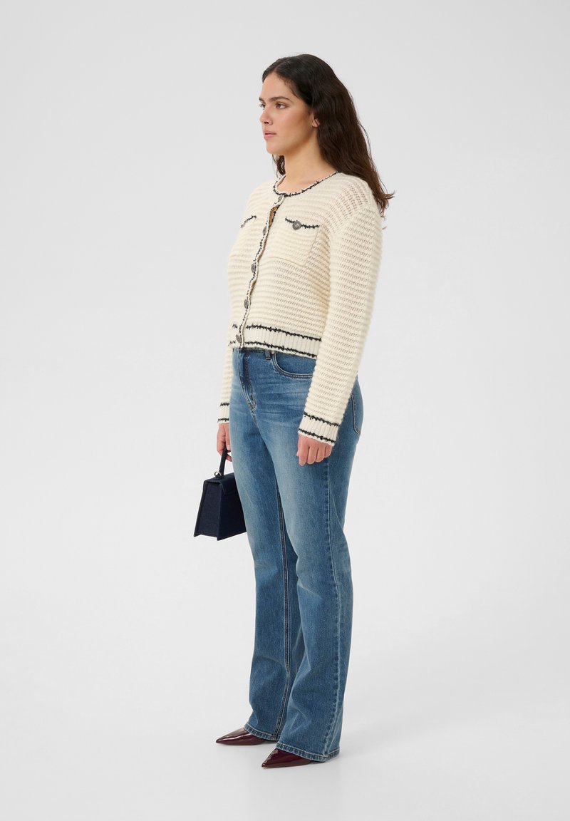 Elfenbeinfarbener Strickcardigan mit schwarzem Besatz und Knöpfen, kombiniert mit blauen Schlagjeans und einer kleinen schwarzen Handtasche. Modell steht seitlich.