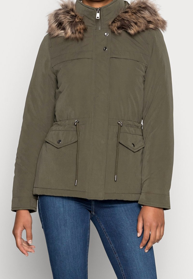 Femme portant une veste d'hiver vert olive avec capuche bordée de fourrure, fermeture éclair à l'avant, boutons-pression et taille avec cordon de serrage, associée à un jean bleu.