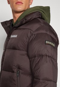 Brun puffjacka med en ficka med dragkedja, logotyp på ärmen och ett överdrag av ett mörkgrönt huva under.