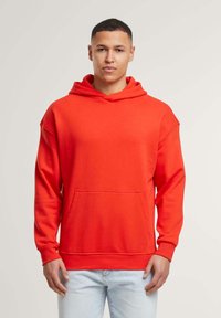 Felpa rossa con cappuccio e tasca a marsupio, tessuto morbido, vestibilità rilassata e cappuccio con cordino, abbinata a jeans azzurri chiari, in piedi contro uno sfondo neutro.