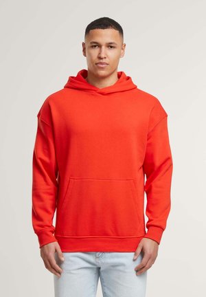 Rød hættetrøje med kængurulomme, blødt stof, afslappet pasform og hætte med snor, parret med lyseblå jeans, stående mod en neutral baggrund.