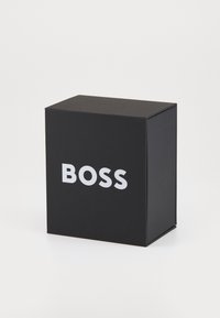Musta kartonkirasia, jossa on matta pinta, etupuolella suuri valkoinen "BOSS"-teksti. Neliönmuotoinen, siisti muotoilu.