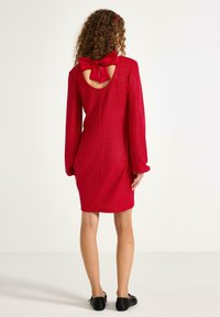 Rotes Glitzerkleid mit langen Ärmeln, offenem Rücken und einer großen roten Schleife, das eng anliegt und oberhalb des Knies endet. Schwarze Ballerinas.