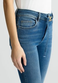 Jeans in denim blu a vita alta con una texture liscia, passanti per la cintura e una tasca frontale. La vestibilità è sartoriale con una leggera elasticità.