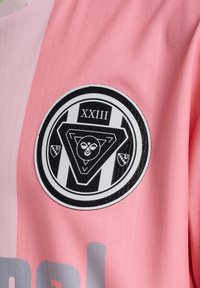 Różowy sportowy strój z teksturowanym czarnym okrągłym logo z geometrycznymi wzorami i napisem "XXIII". Szare litery widnieją poniżej logo.