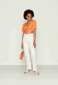 Top cropped laranja atado, jeans brancos de cintura alta, sapatos slip-on de cor clara e bolsa laranja com alça de corrente. Acessórios: óculos de sol pequenos.