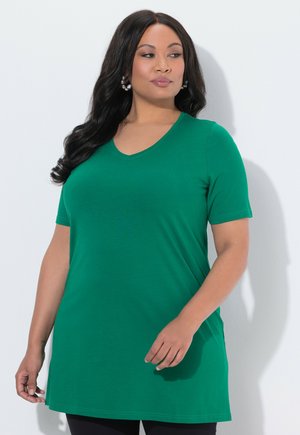 Frau mit langen dunklen Haaren, die eine grüne kurzärmlige Tunika mit V-Ausschnitt trägt, steht vor weißem Hintergrund und schaut zur Seite.