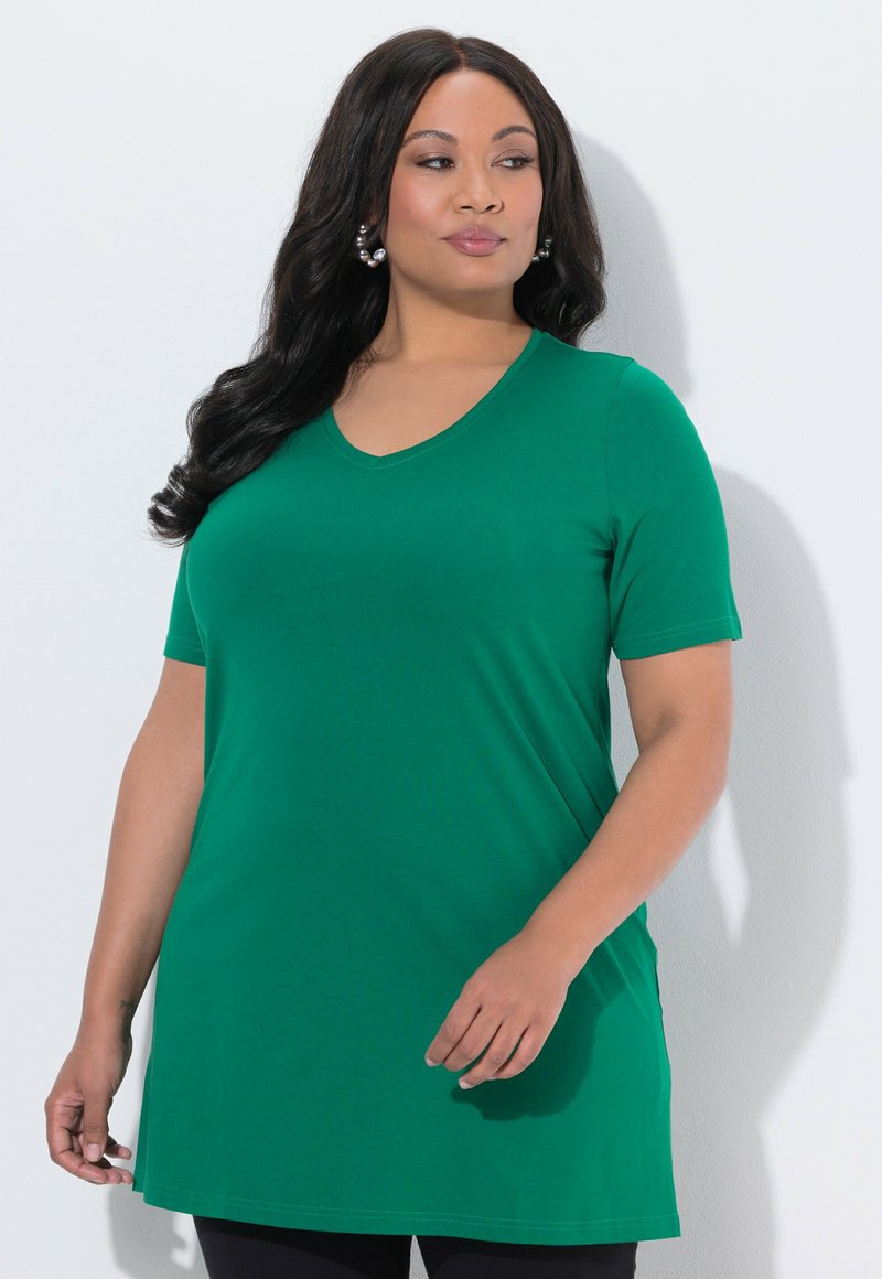 Vrouw met lang donker haar, gekleed in een groen tuniek met korte mouwen en een V-hals, staand tegen een witte achtergrond, kijkend naar de zijkant.