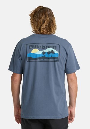 Mann mit schulterlangen Haaren, der ein blaues Billabong-T-Shirt mit tropischem Sonnenuntergang und Palme auf dem Rücken trägt.