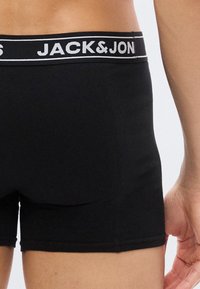 Αρσενικός κορμός που φοράει μαύρα εσώρουχα Jack & Jones με λωρίδα μέσης με λογότυπο, εμφανίζεται από πίσω με το δεξί χέρι ορατό.