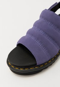 Lilla semsket sandaler med en bred, polstret stropp, åpen tå og sort gummisåle med gule stingdetaljer.