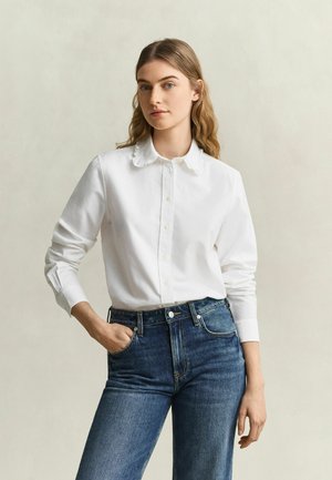 Junge Frau mit langem welligem Haar trägt ein weißes Hemd mit Knöpfen und Kragen und blaue hoch taillierte Jeans, steht mit einer Hand in der Tasche.