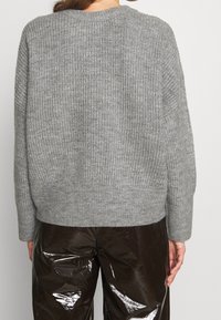 Personne portant un pull gris côtelé en tricot et un pantalon en vinyle brun foncé brillant, tourné de dos face à un fond uni.