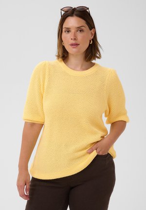 Frau mit schulterlangem braunem Haar, die ein gelbes Strickoberteil, dunkle Hose, goldene Creolen und eine Sonnenbrille auf dem Kopf trägt.