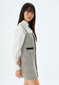 Vestido a cuadros con un patrón en blanco y negro, con un diseño ajustado, tirantes ajustables, botones dorados y bolsillos laterales.