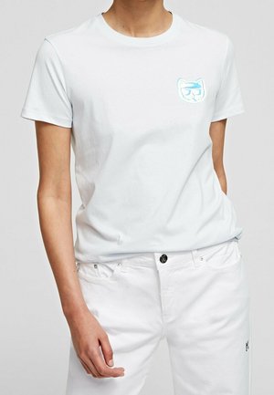 Persona con una camiseta blanca lisa de manga corta con un pequeño logo azul de cara de gato en el pecho izquierdo y pantalones blancos con un botón negro.