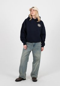 Tânără purtând o șapcă bej, hanorac bleumarin cu logo, blugi largi albastru deschis și încălțăminte maro închis, stând în fața unui fundal alb.