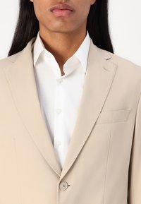 Blazer beige con una texture liscia, rever affilati e chiusura a un bottone. Dettaglio della tasca inserita, indossato sopra una camicia bianca con bottoni.