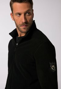 Chaqueta de forro polar negra con cierre de cremallera en el frente, cuello alto y parche de águila bordado en la manga izquierda. Textura suave, diseño ajustado.