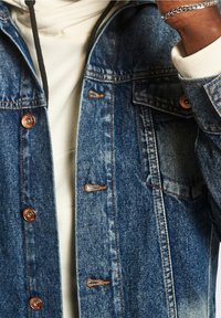 Giubbotto in denim blu scuro con accessori in rame e cuciture a contrasto. Presenta due tasche sul petto e una vestibilità morbida sopra un leggero hoodie.