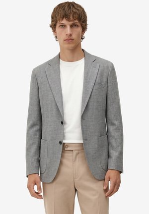 Junger Mann trägt einen hellgrauen Blazer, weißes T-Shirt mit Rundhalsausschnitt und beige Hose, steht vor einem schlichten hellen Hintergrund.