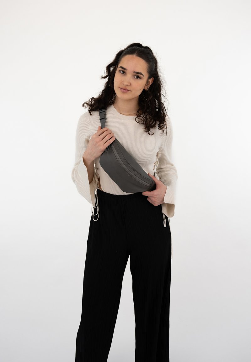 Borsa a tracolla grigia realizzata in tessuto liscio con una cerniera diagonale, indossata attraversato il corpo; abbinata a un maglione leggero e pantaloni neri plissettati.