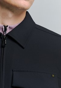 Zwarte jas met een ritssluiting aan de voorkant en kraag, gemaakt van een gestructureerde stof. Bevat een borstzak met een logo accent. Stevige structuur.