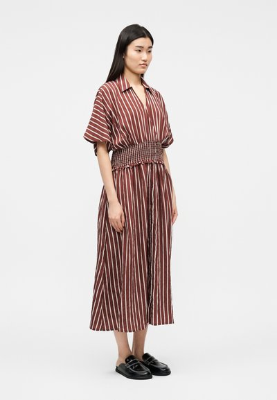 Γυναίκα που φοράει φόρεμα με καφέ και λευκές κάθετες ρίγες, με κοντά μανίκια και φαρδιά, υφασμάτινη ζώνη στη μέση, συνδυασμένο με μαύρα παπούτσια τύπου slip-on.